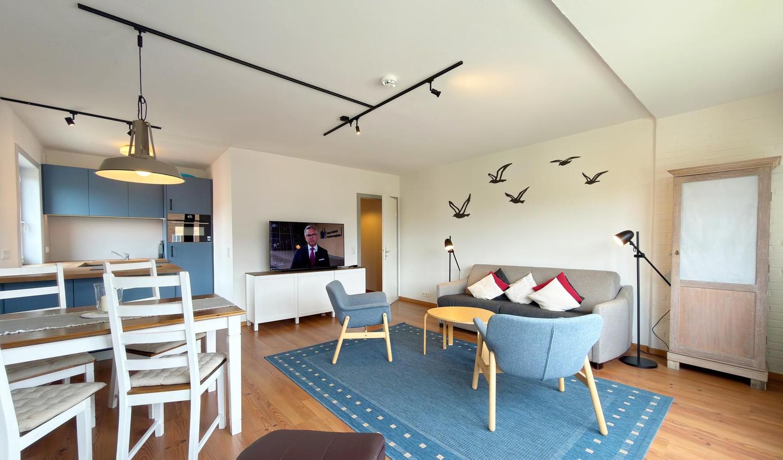 Wohnbereich mit Sofa, zwei Stühlen, TV und Küchenbereich. Wanddekoration mit Vögeln.
