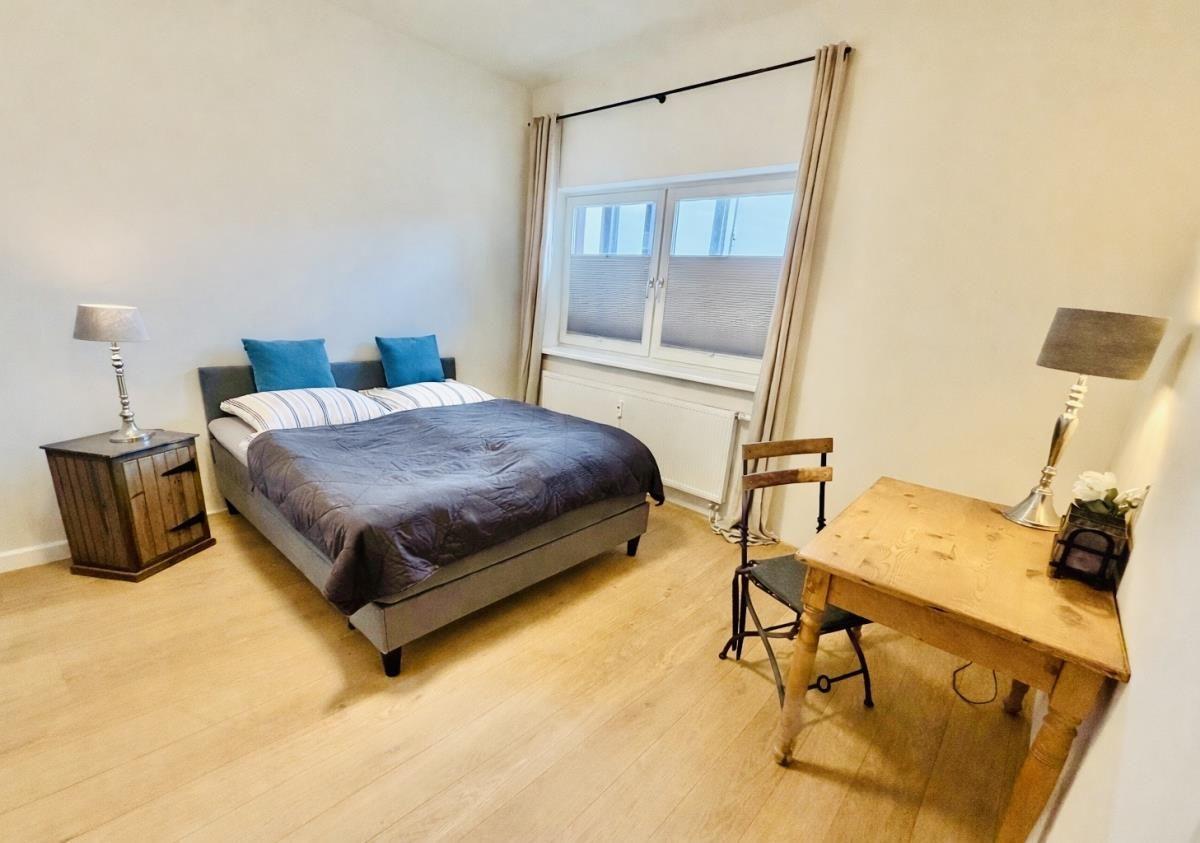 Schlafzimmer mit Bett, Schreibtisch und Fenster. Holzboden und Nachttische mit Lampen.