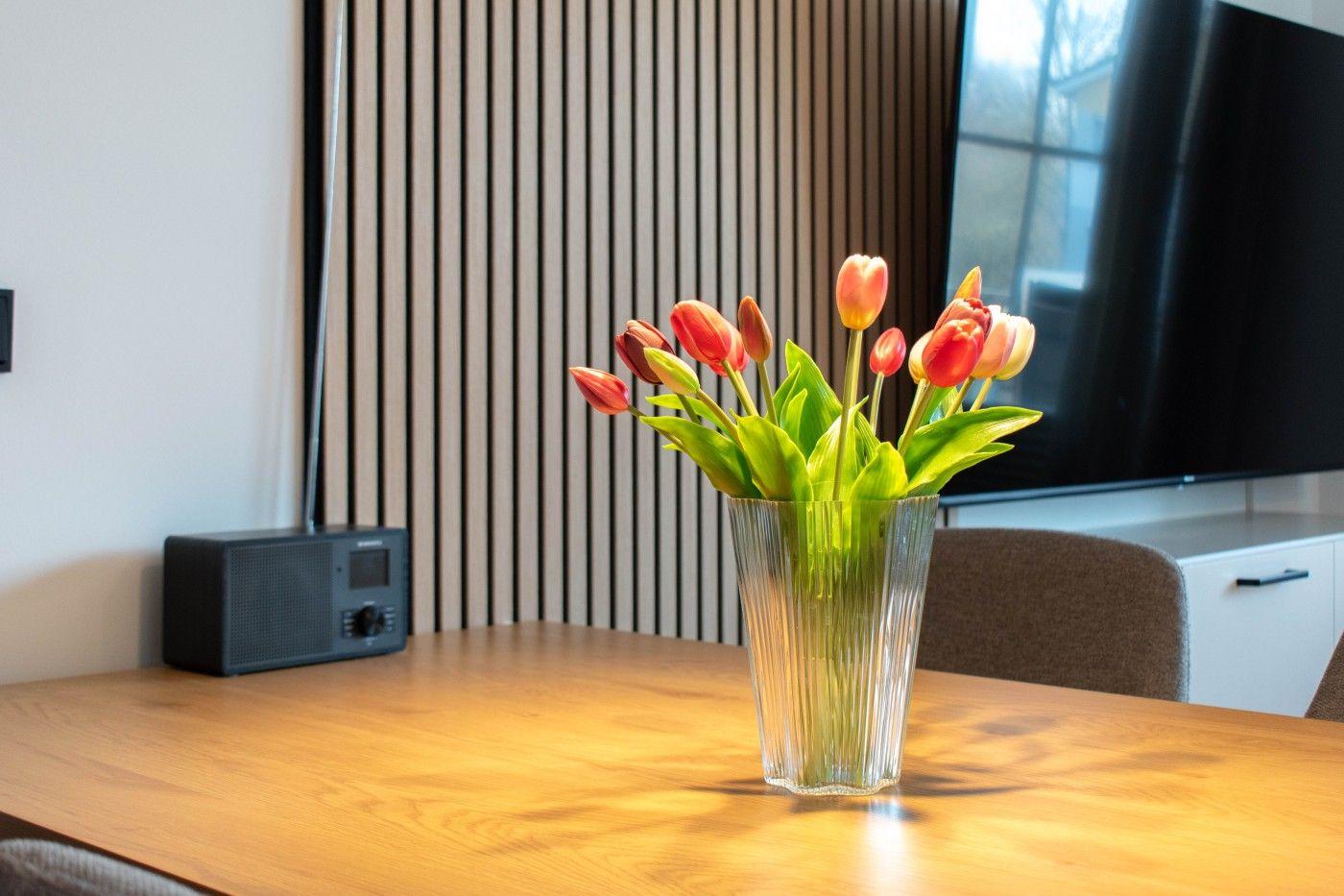Ein Glas mit Tulpen steht auf einem Holztisch. Im Hintergrund ist ein Fernseher und eine Wandverkleidung zu sehen.