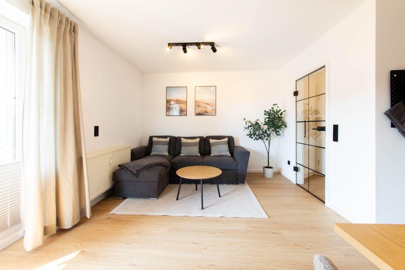 Wohnzimmer mit Sofa, Tisch und Pflanze. Holzboden und Fenster mit Vorhängen.