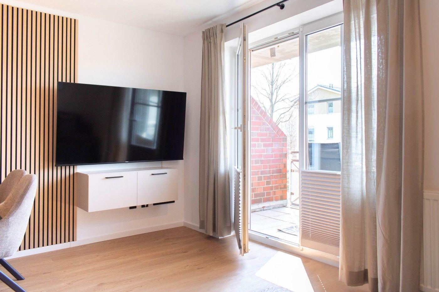Wohnzimmer mit Fernseher, Holzboden und Balkontür.
