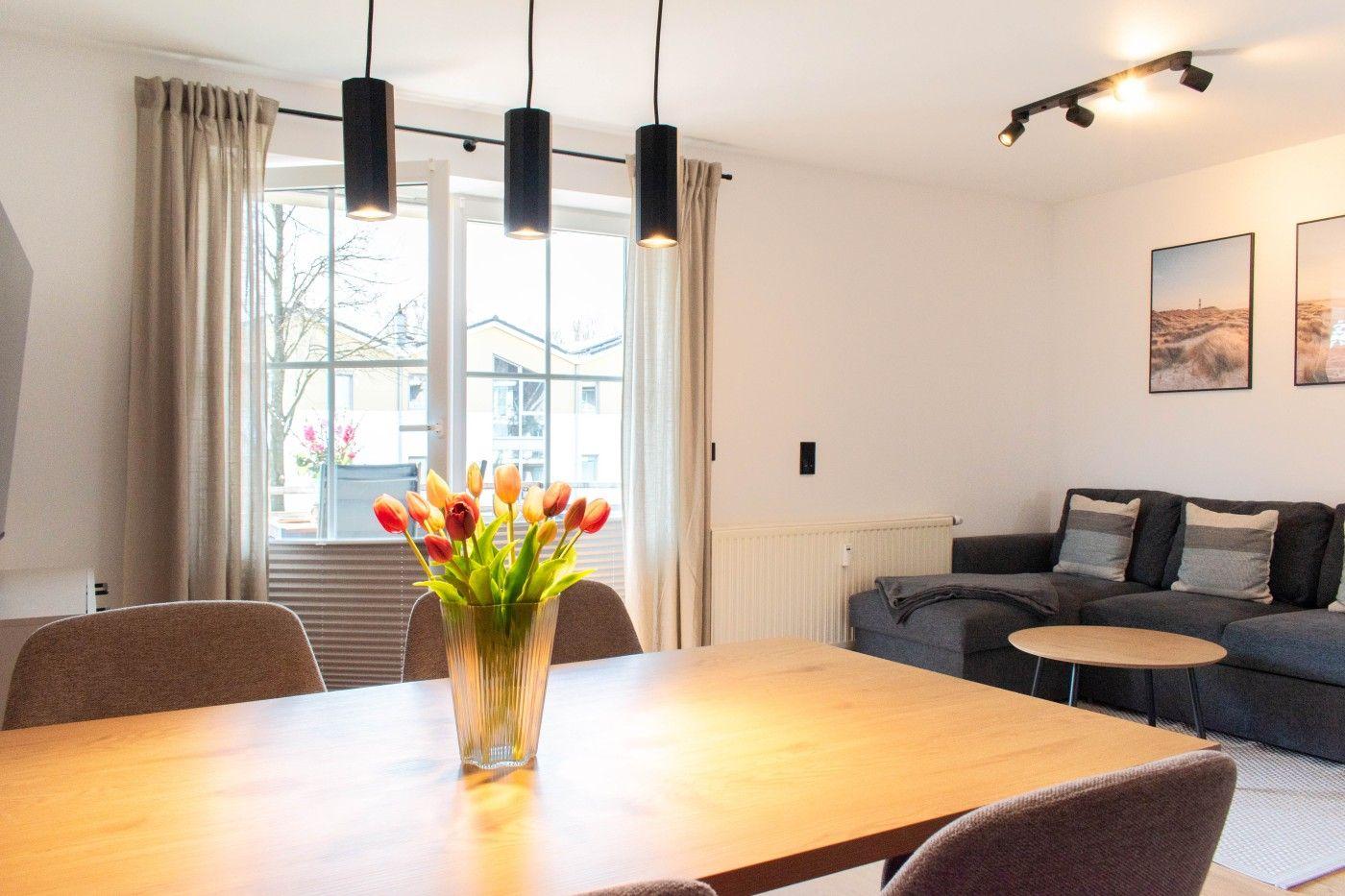 Essbereich mit Tisch, Stühlen und Blumen. Wohnbereich mit Sofa und Wandbildern.
