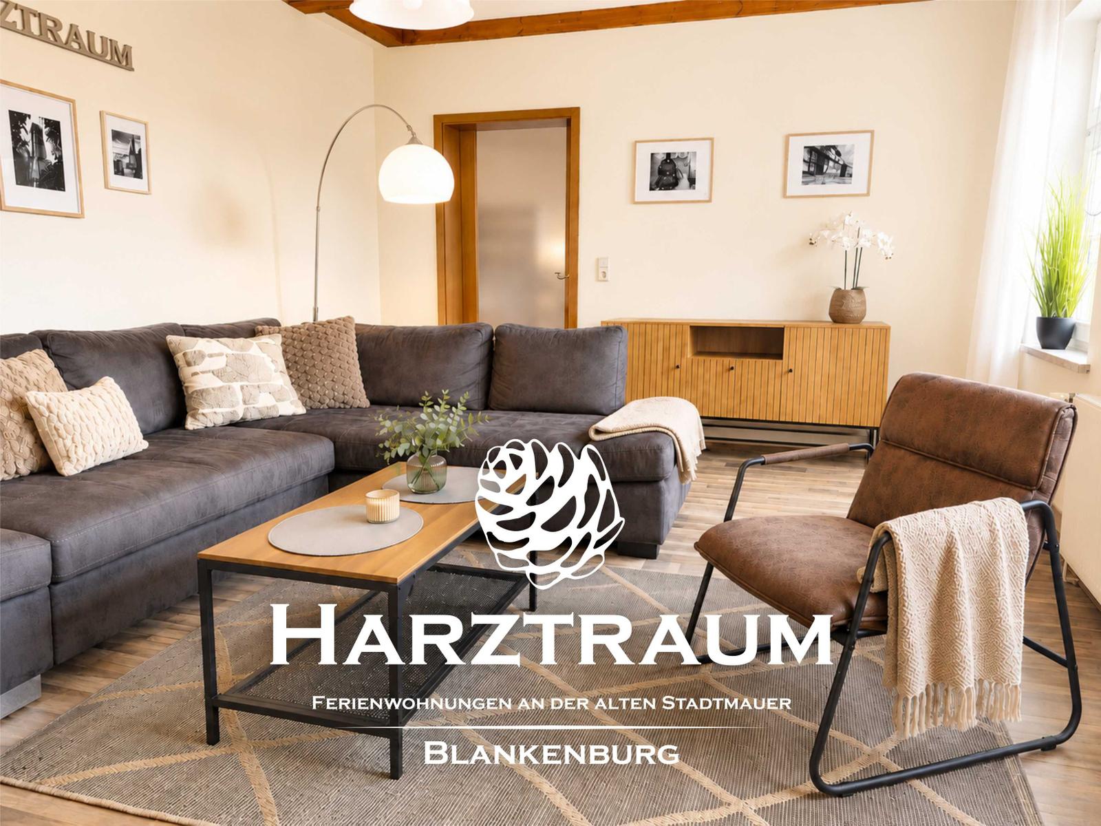 Wohnzimmer mit grauem Sofa, Holztisch und brauner Sessel. Wandbild und Pflanzen sichtbar.