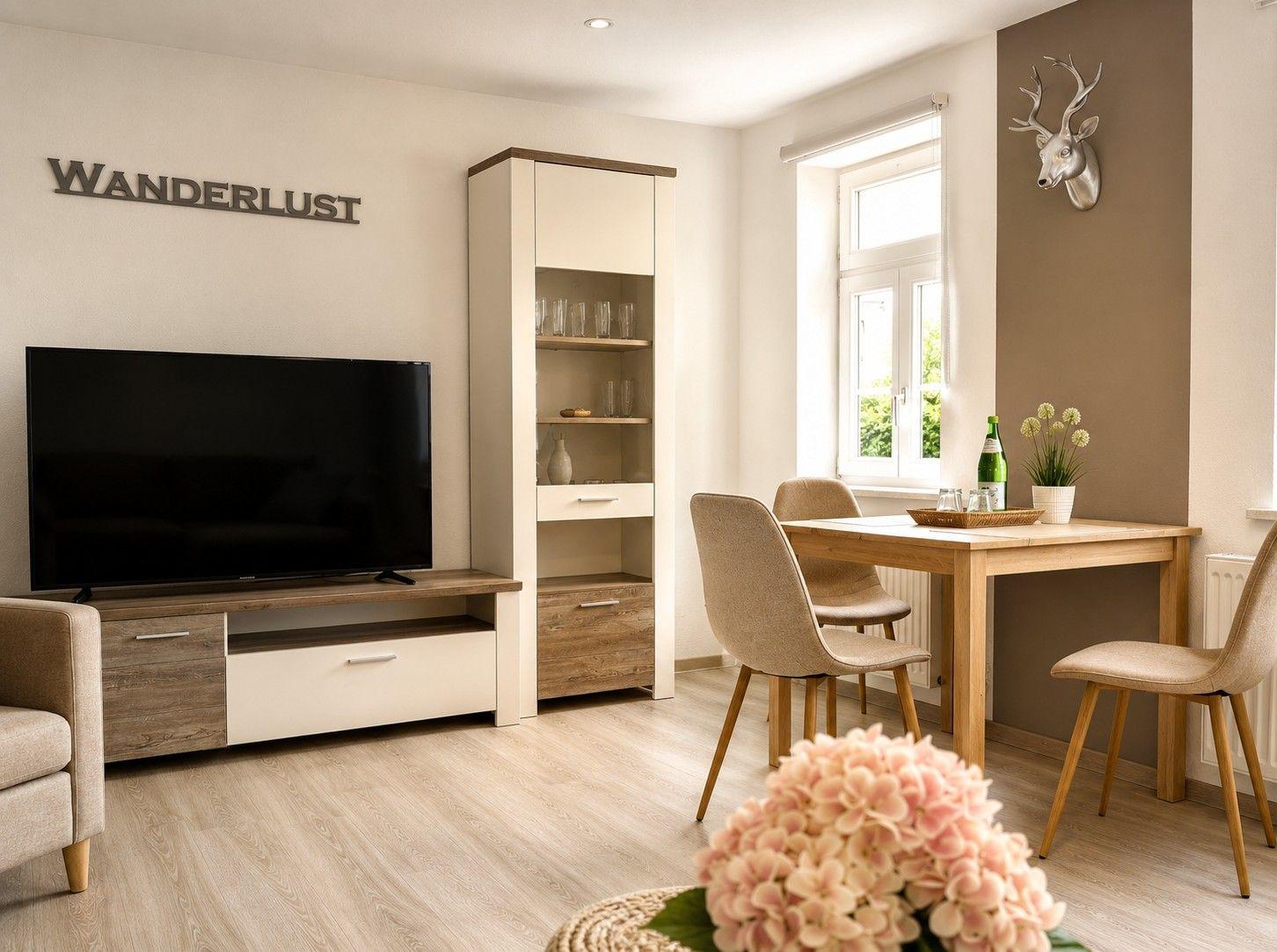 Wohn- und Essbereich mit TV, Tisch und Stühlen. Wanddekoration mit Hirsch und Schriftzug.
