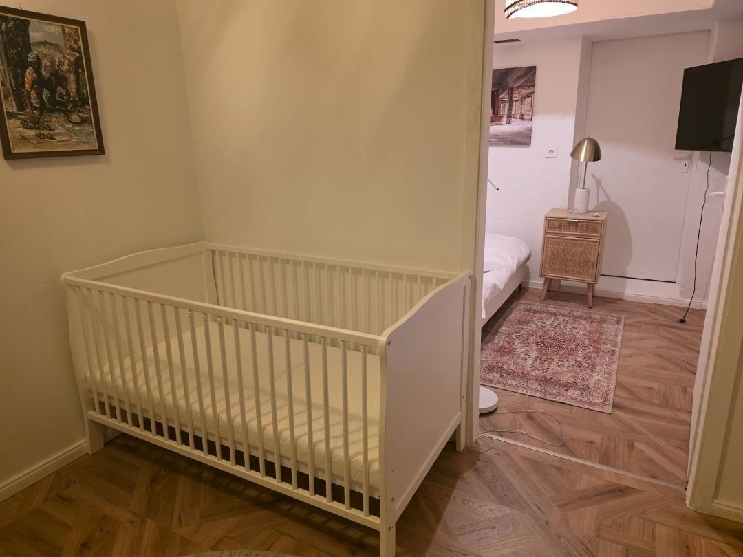 Ein weißes Babybett steht im Vordergrund. Im Hintergrund ist ein Schlafzimmer mit einem Bett und einem Nachttisch.