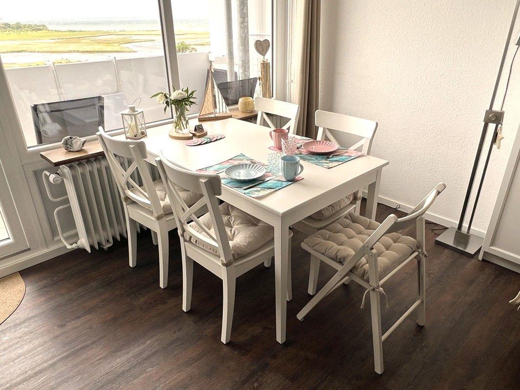 Dining area with white table and chairs, view of landscape through large windows.