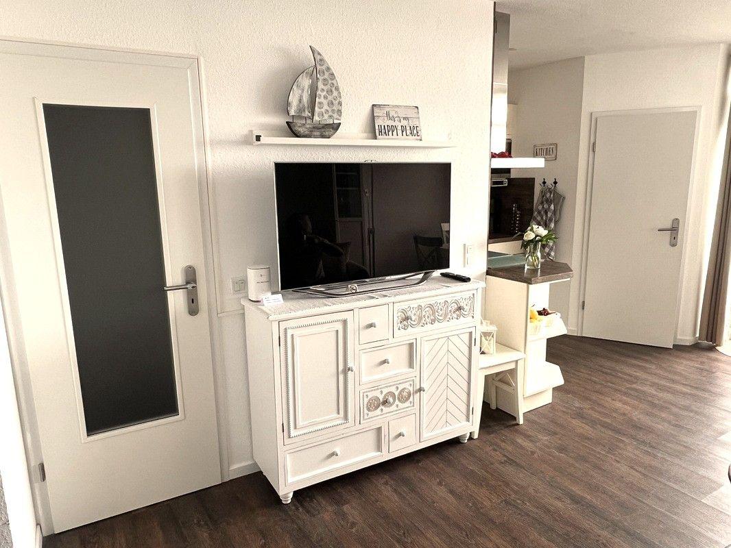 Living room with TV cabinet, door, and kitchen area in background.