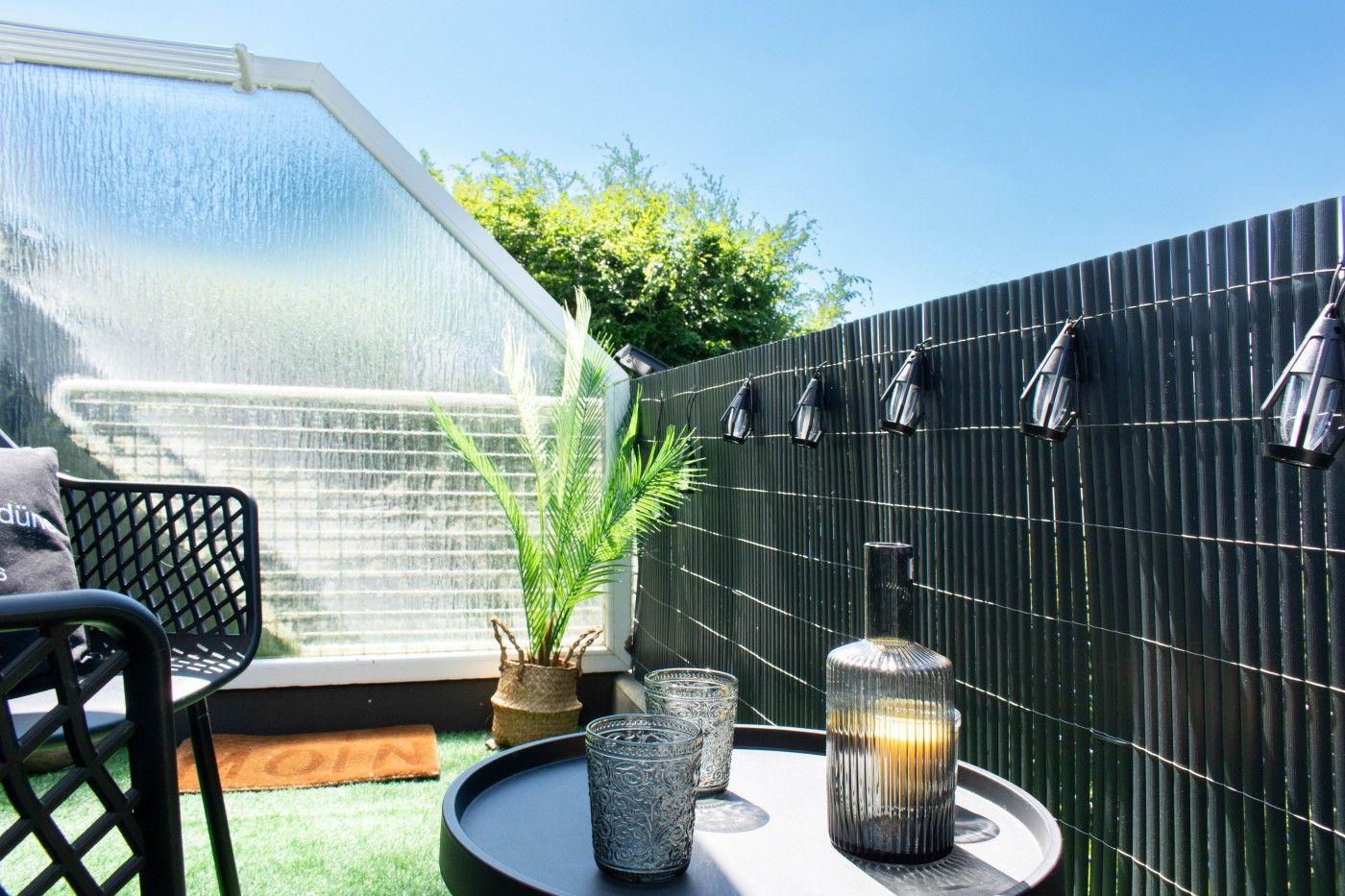 Terrasse mit Tisch, Stuhl, Pflanze und Lichterkette an schwarzem Zaun.