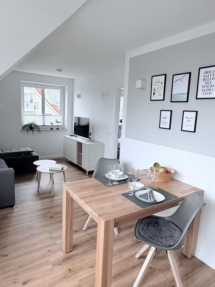 Essbereich mit Holztisch und Stühlen. Wand mit Bildern. Wohnbereich mit TV und Fenster.