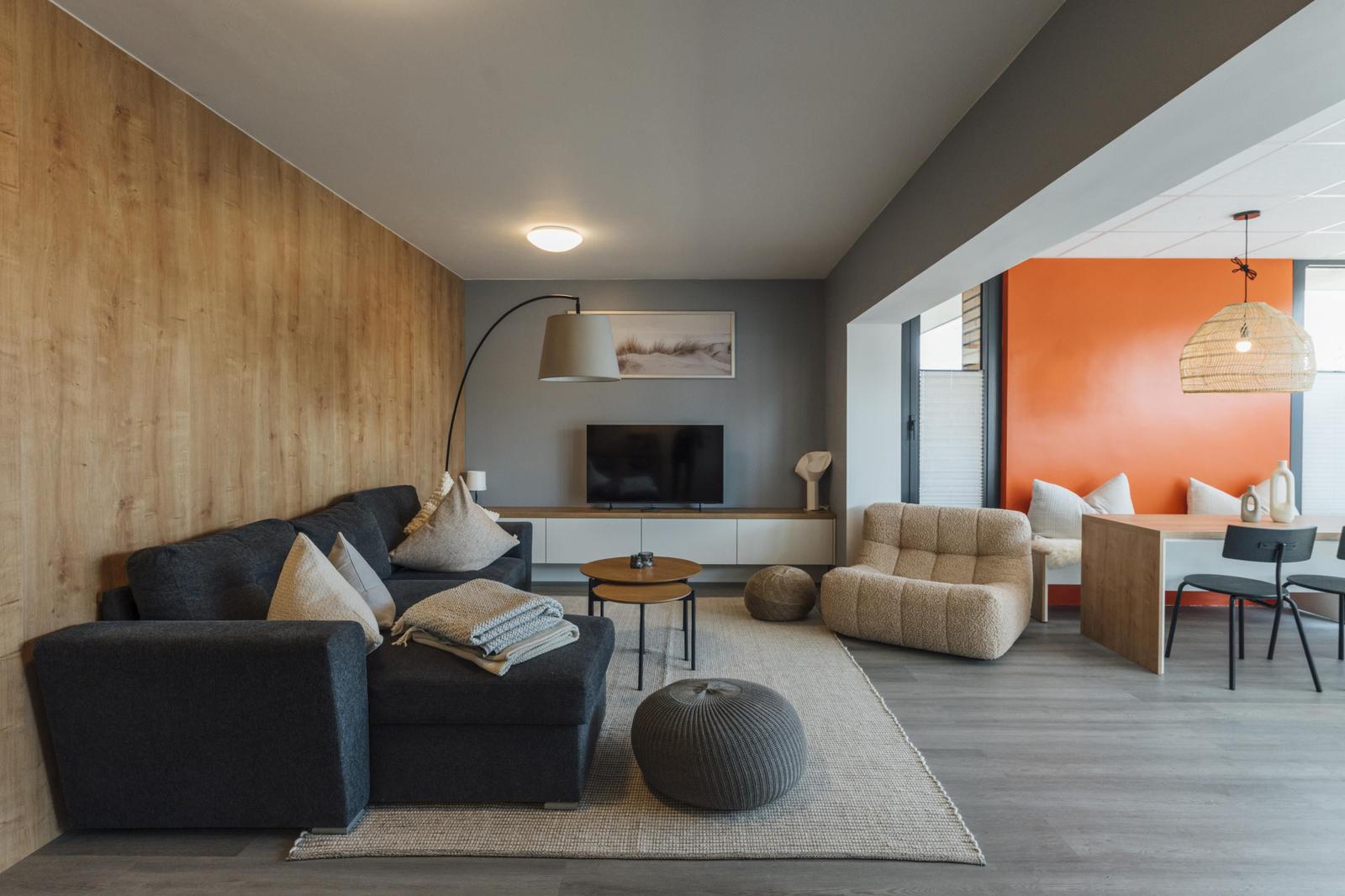 Wohnzimmer mit Sofa, Sessel und Esstisch. Holzwand und orange Wand.