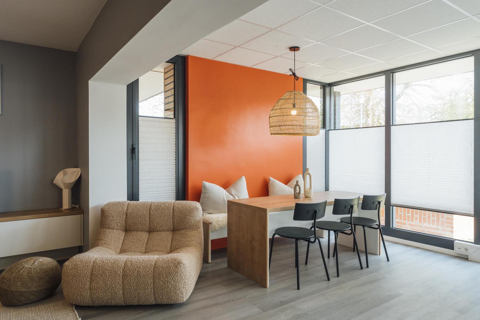 Ein Raum mit Essbereich, Sofa und orange Wand. Große Fenster und hängende Lampe.