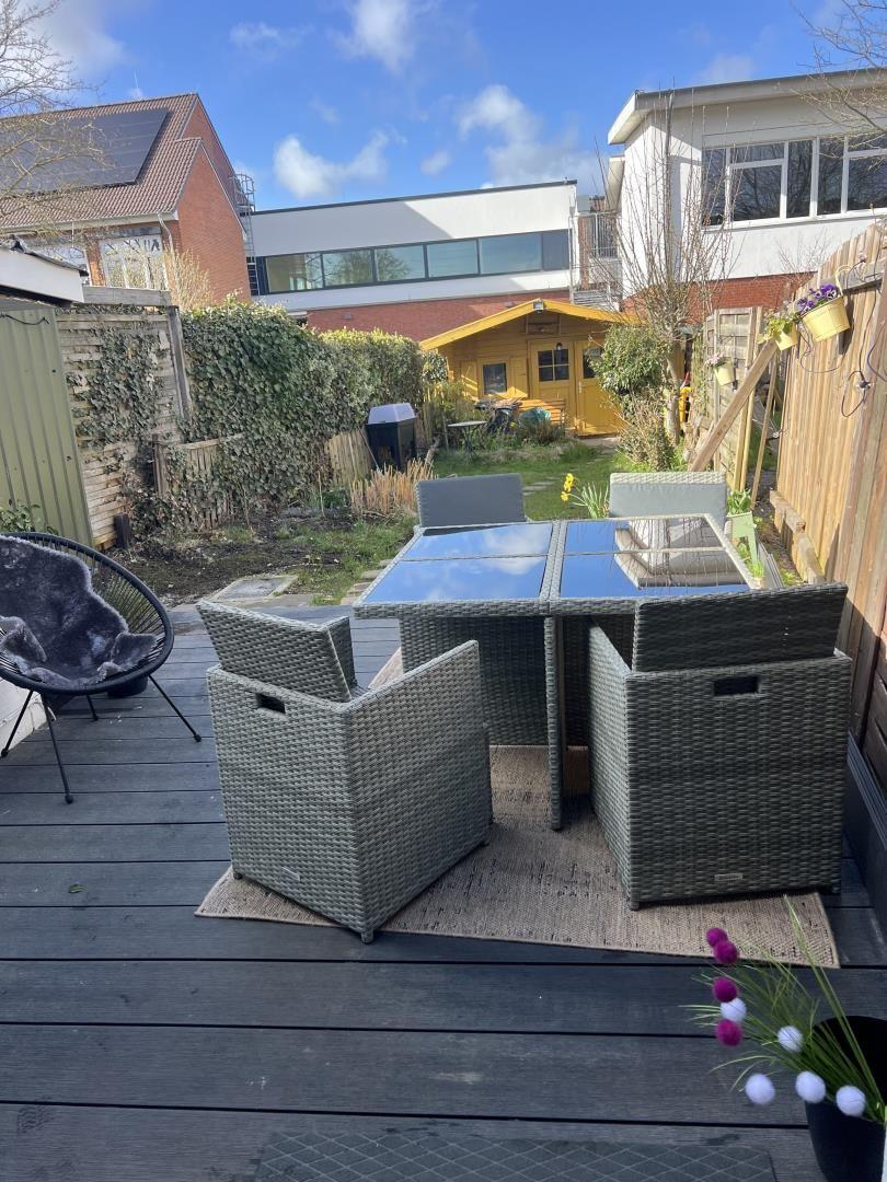 Terrasse mit Rattanmöbeln, Tisch und Sitzgelegenheiten. Hinterhof mit Garten und gelbem Holzhaus.