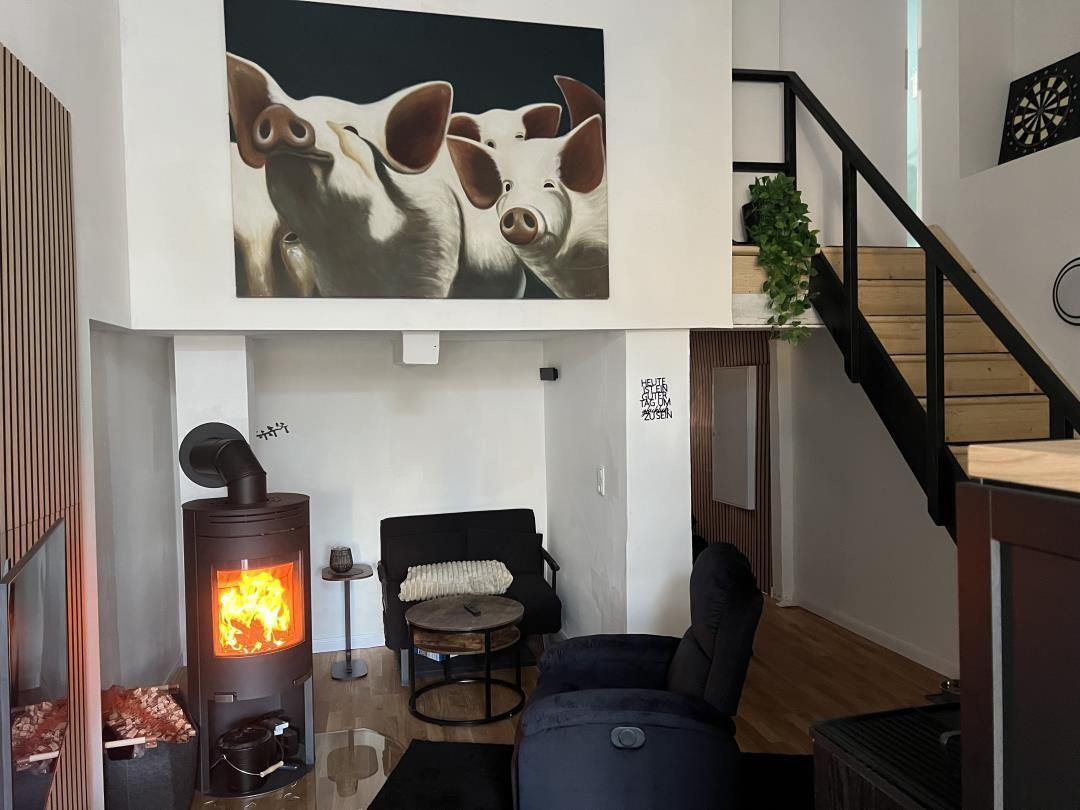 Wohnbereich mit Kamin, Sitzgelegenheiten und Treppenaufgang. Wandbild zeigt Schweine.