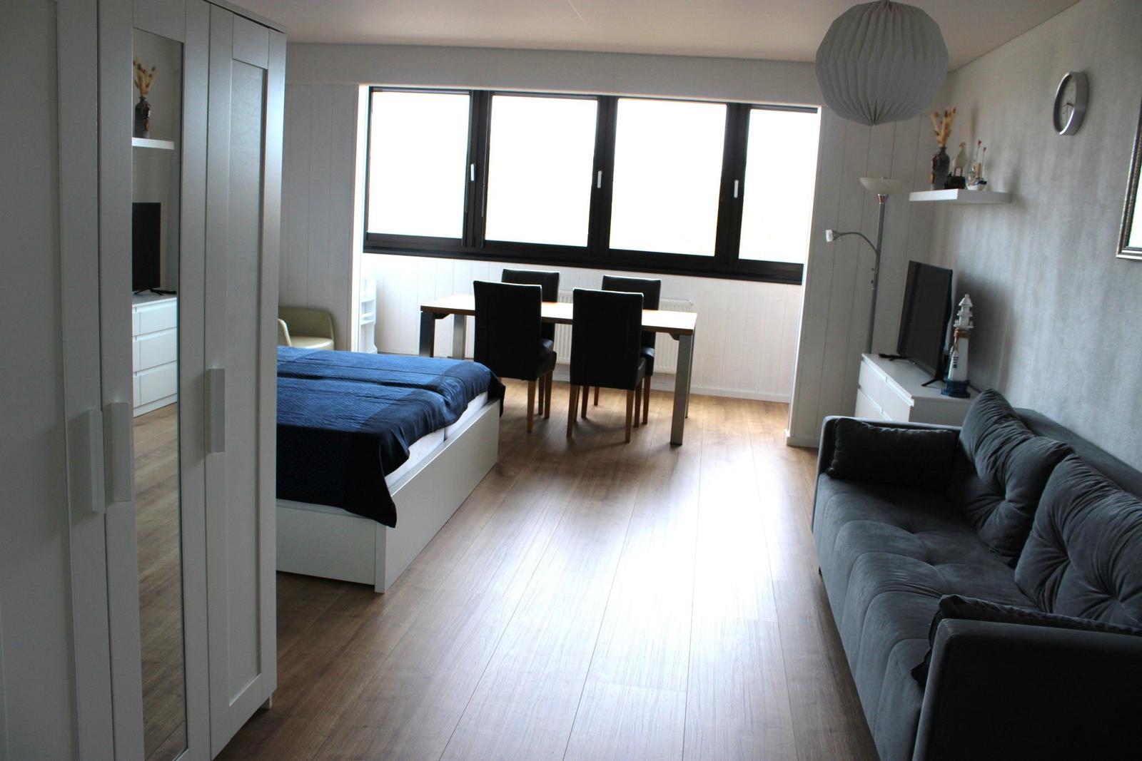 Wohnraum mit Sofa, Schlafbereich und Esstisch. Große Fenster und Holzboden.