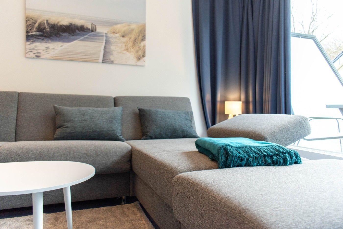 Ein grauer Ecksofa mit Kissen und einer Decke vor einem Fenster mit Vorhängen.