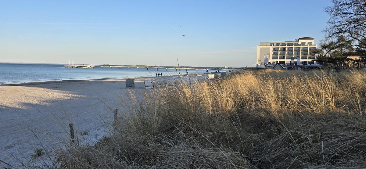 Strand mit Dünen, Steg und Hotel im Hintergrund.