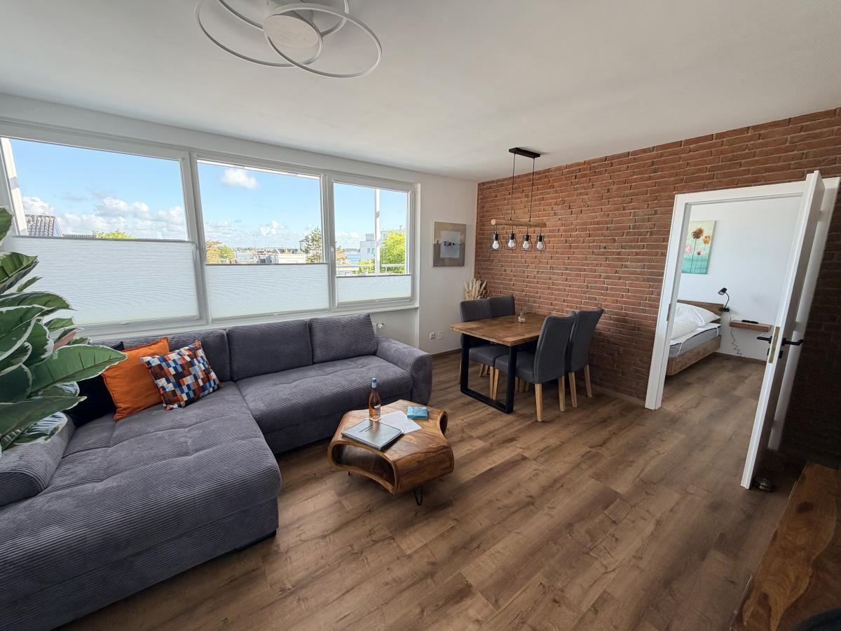 Wohnzimmer mit Sofa, Esstisch und Schlafzimmer im Hintergrund. Holzboden und Backsteinwand.