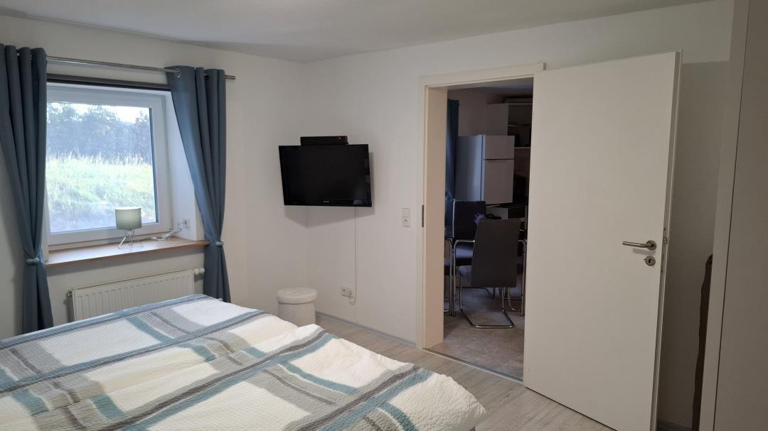 Schlafzimmer mit Bett, TV und Fenster. Tür führt in die Küche.
