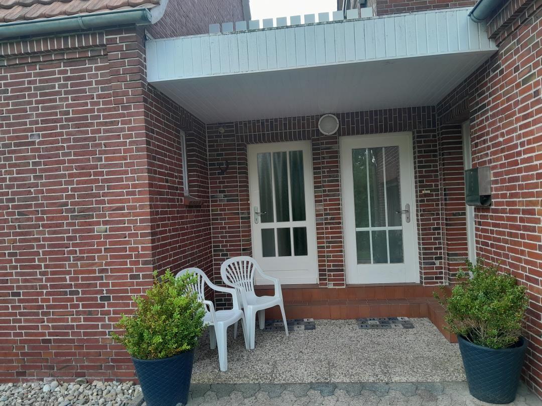 Eingangsbereich mit zwei weißen Stühlen und Pflanzen in blauen Töpfen vor rotem Backsteinhaus.
