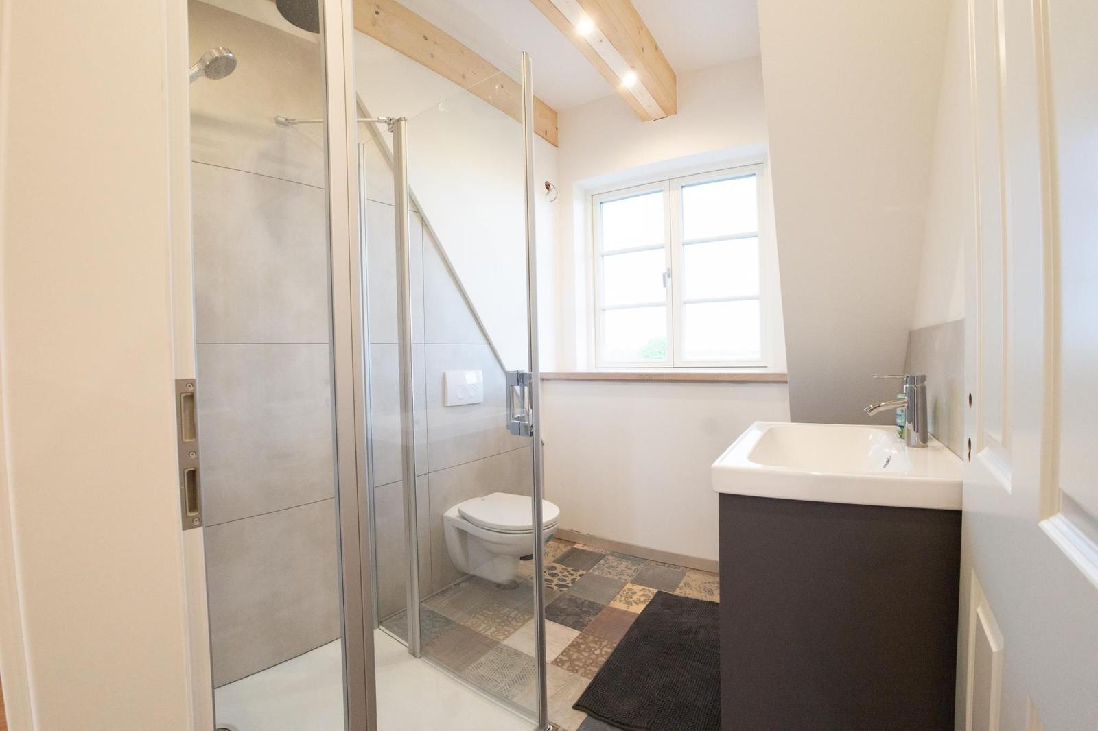 Badezimmer mit Dusche, Toilette und Waschbecken. Holzbalken und Fenster sichtbar.