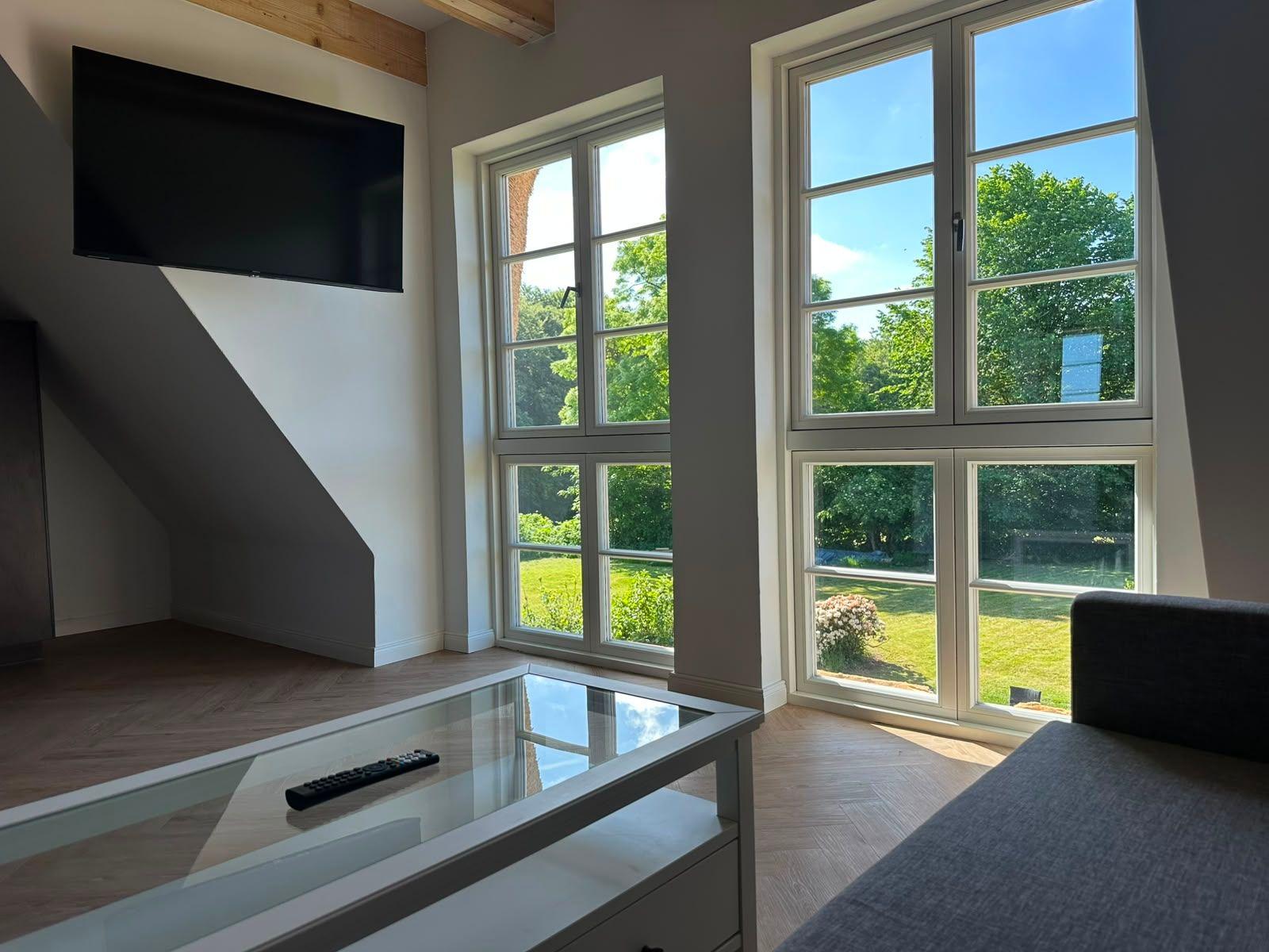 Wohnraum mit großen Fenstern, TV und Glas-Tisch. Blick auf grünen Garten.