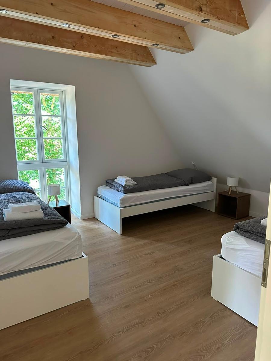 Zimmer mit drei Betten, Holzdecken und Fenster mit Blick auf Grün.