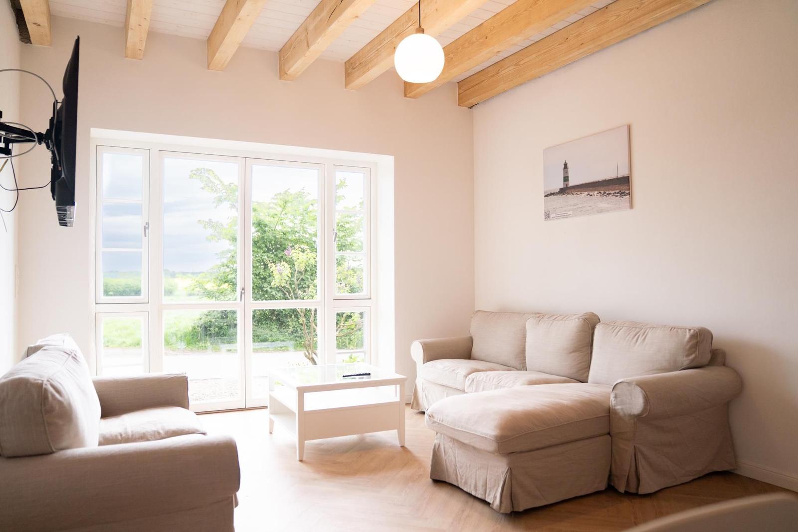Wohnzimmer mit Sofa, Sessel und Glas-Tisch. Fenster mit Blick auf Garten. Holzbalken an der Decke.