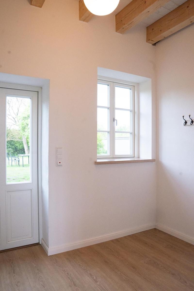 Raum mit Holzfußboden, weißen Wänden, Fenster und Tür. Holzbalken an der Decke.