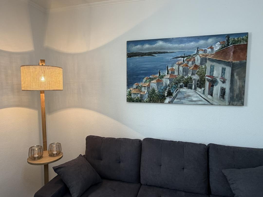 A sofa with cushions, a floor lamp, and a wall painting of coastal houses.