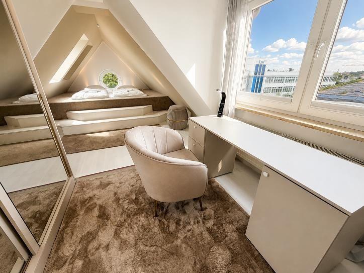 Schlafzimmer mit Dachschrägen, Bett, Schreibtisch und Sessel. Blick auf Stadt durch Fenster.