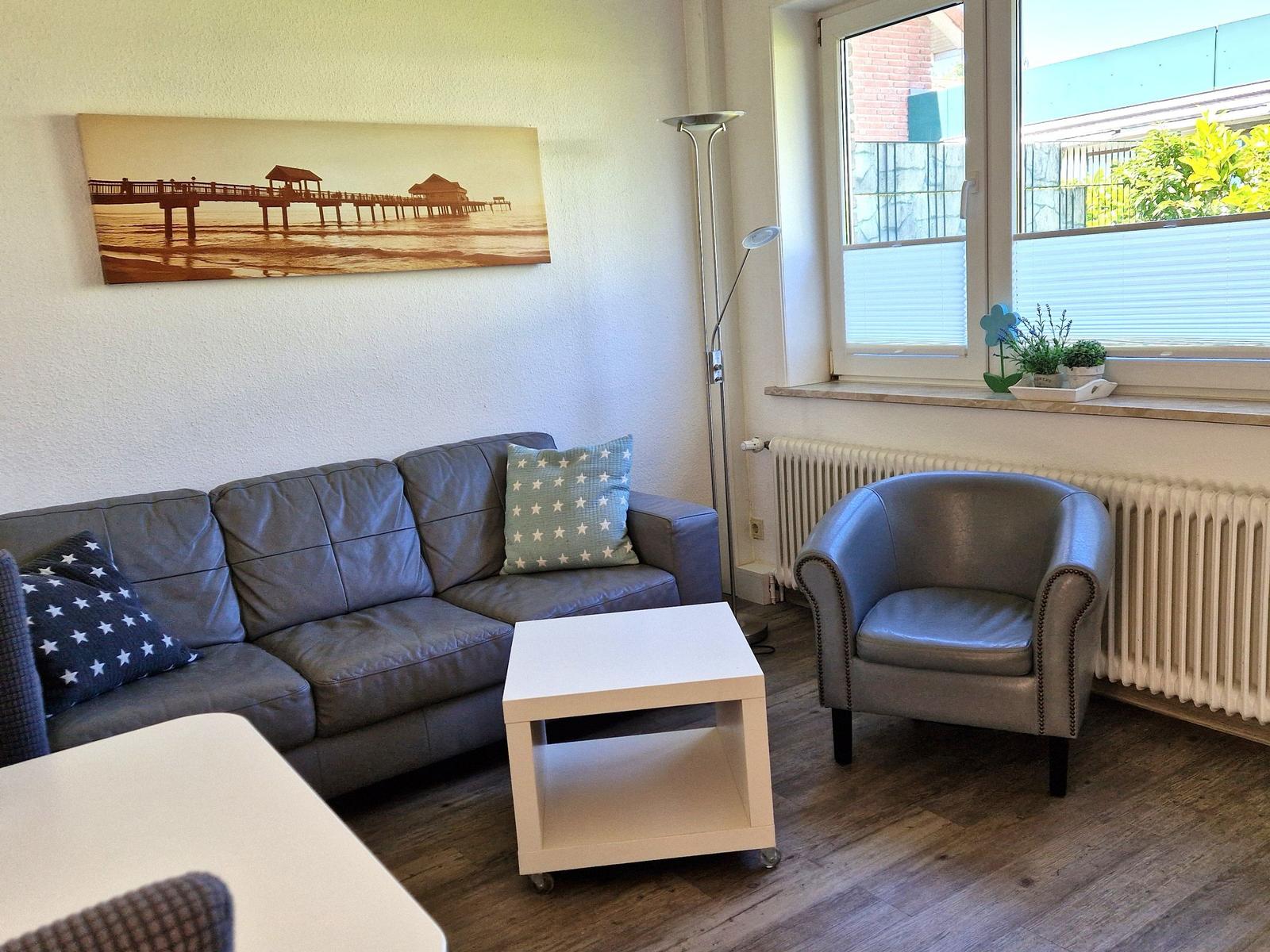 Wohnzimmer mit Sofa, Sessel und Tisch. Wandbild zeigt Pier. Fenster mit Pflanzen.