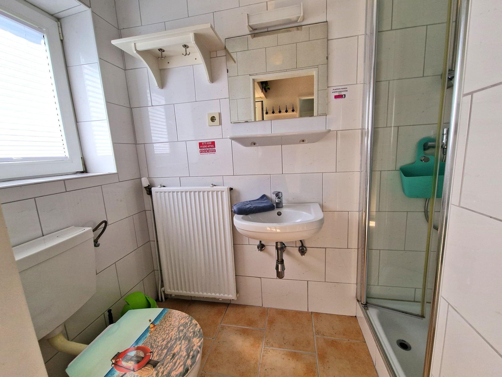 Badezimmer mit Dusche, Waschbecken und Toilette. Weiße Fliesen und Fenster mit Jalousie.
