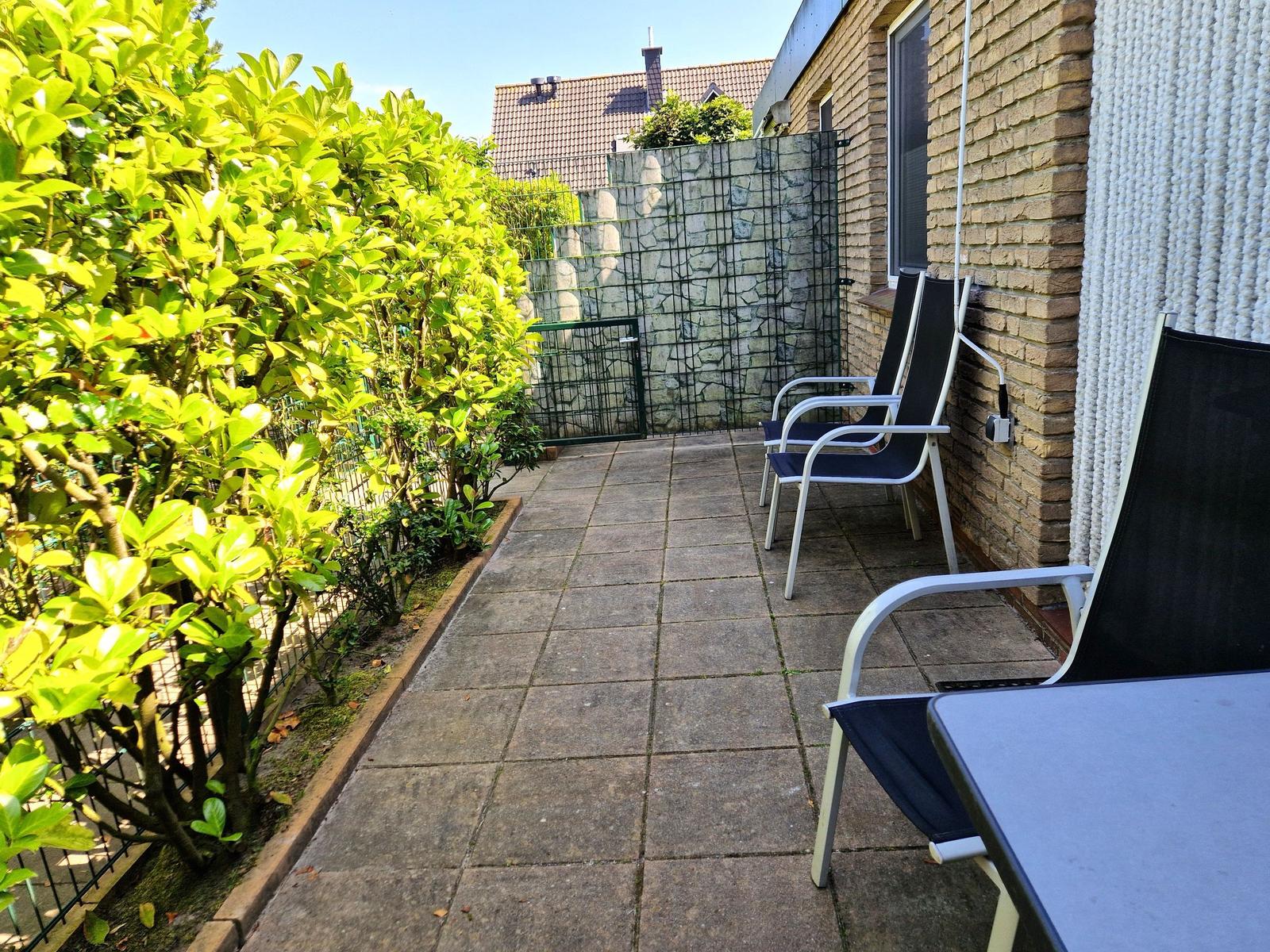Terrasse mit Sitzgelegenheiten und grüner Hecke. Steinmauer und Fenster sind sichtbar.