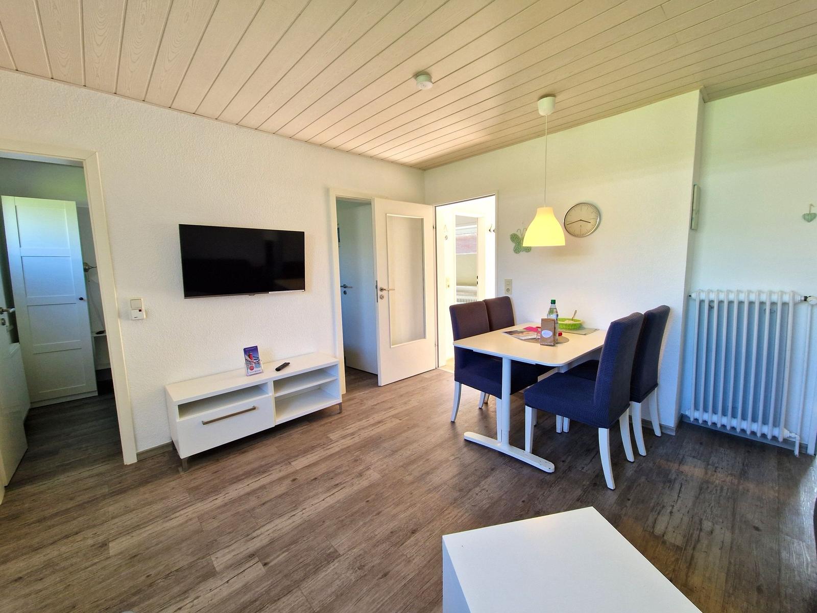 Wohn- und Essbereich mit TV, Tisch und Stühlen sowie Holzboden und weißen Wänden.