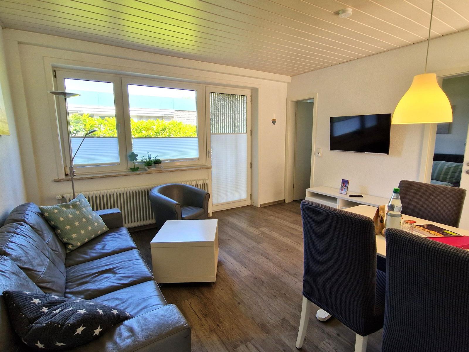 Wohnzimmer mit Sofa, Esstisch und Fernseher. Holzboden und Fenster mit Blick.