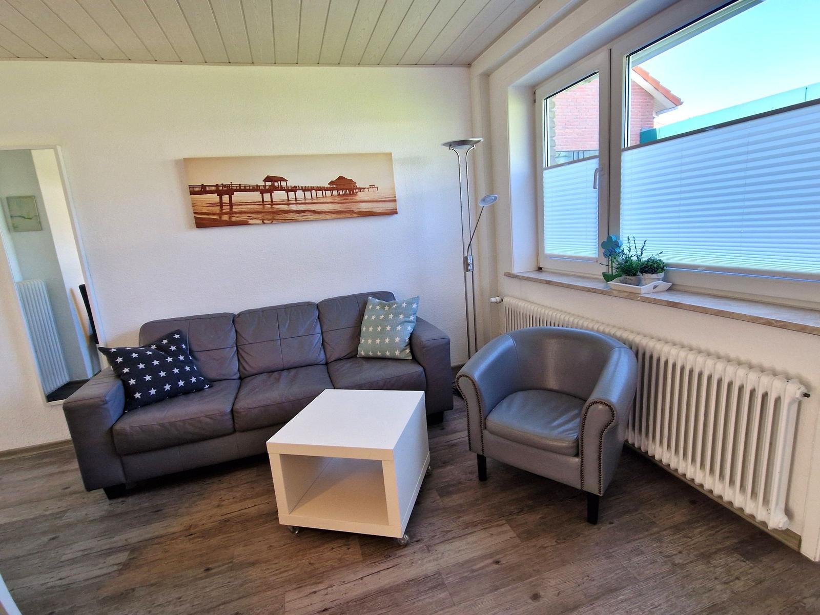 Wohnzimmer mit Sofa, Sessel und Couchtisch. An der Wand hängt ein Bild einer Promenade.