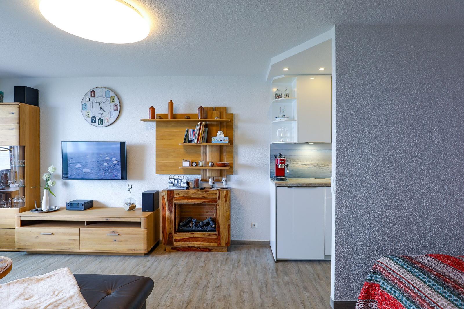 Wohnbereich mit Fernseher, Holzregal und Kamin. Küche mit roter Kaffeemaschine.