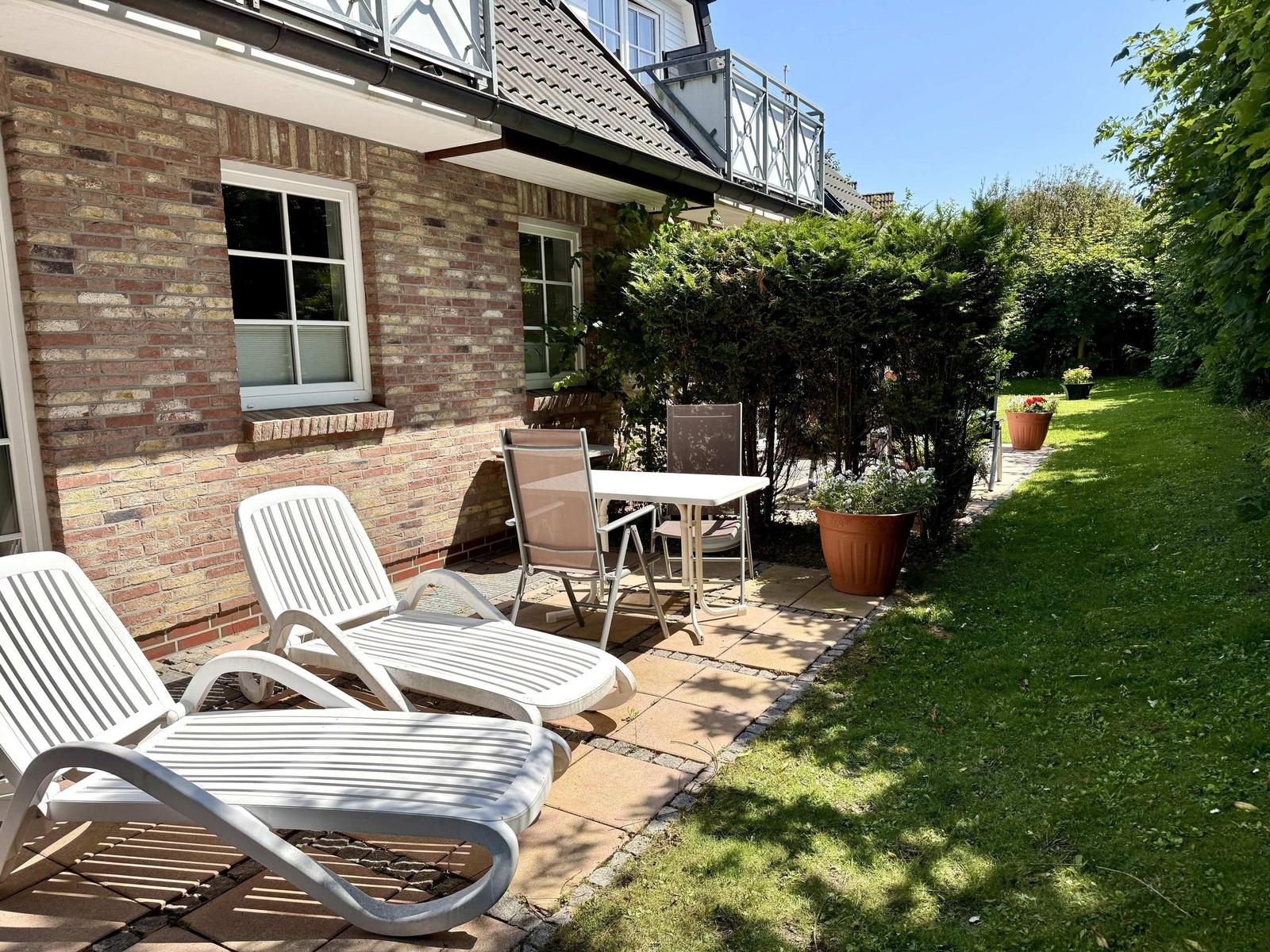 Terrasse mit Liegestühlen, Tisch und Stühlen. Grünfläche und Pflanzen im Hintergrund.