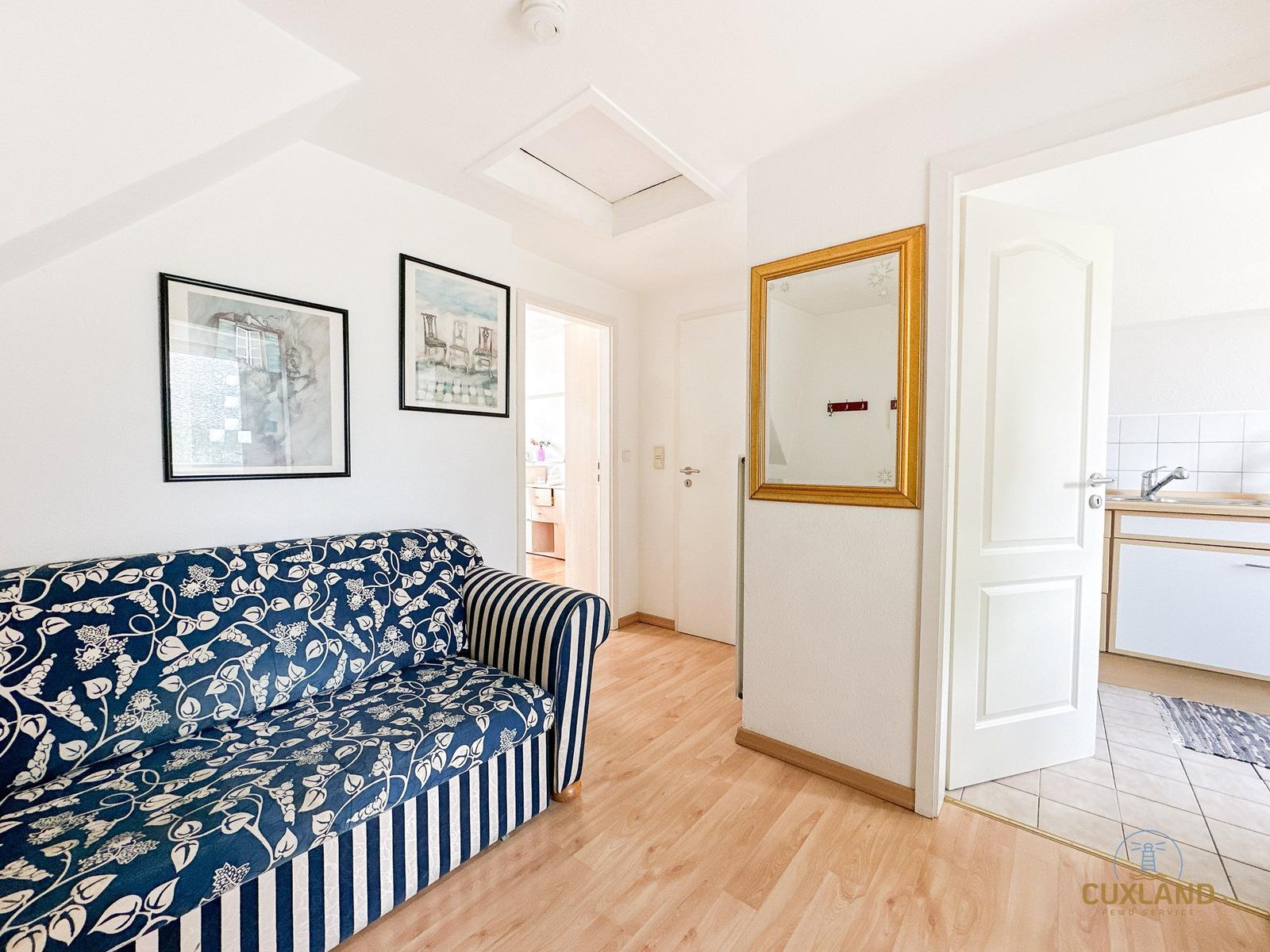 Wohnzimmer mit Sofa, Wandbildern und Holzboden. Tür führt in Küche.