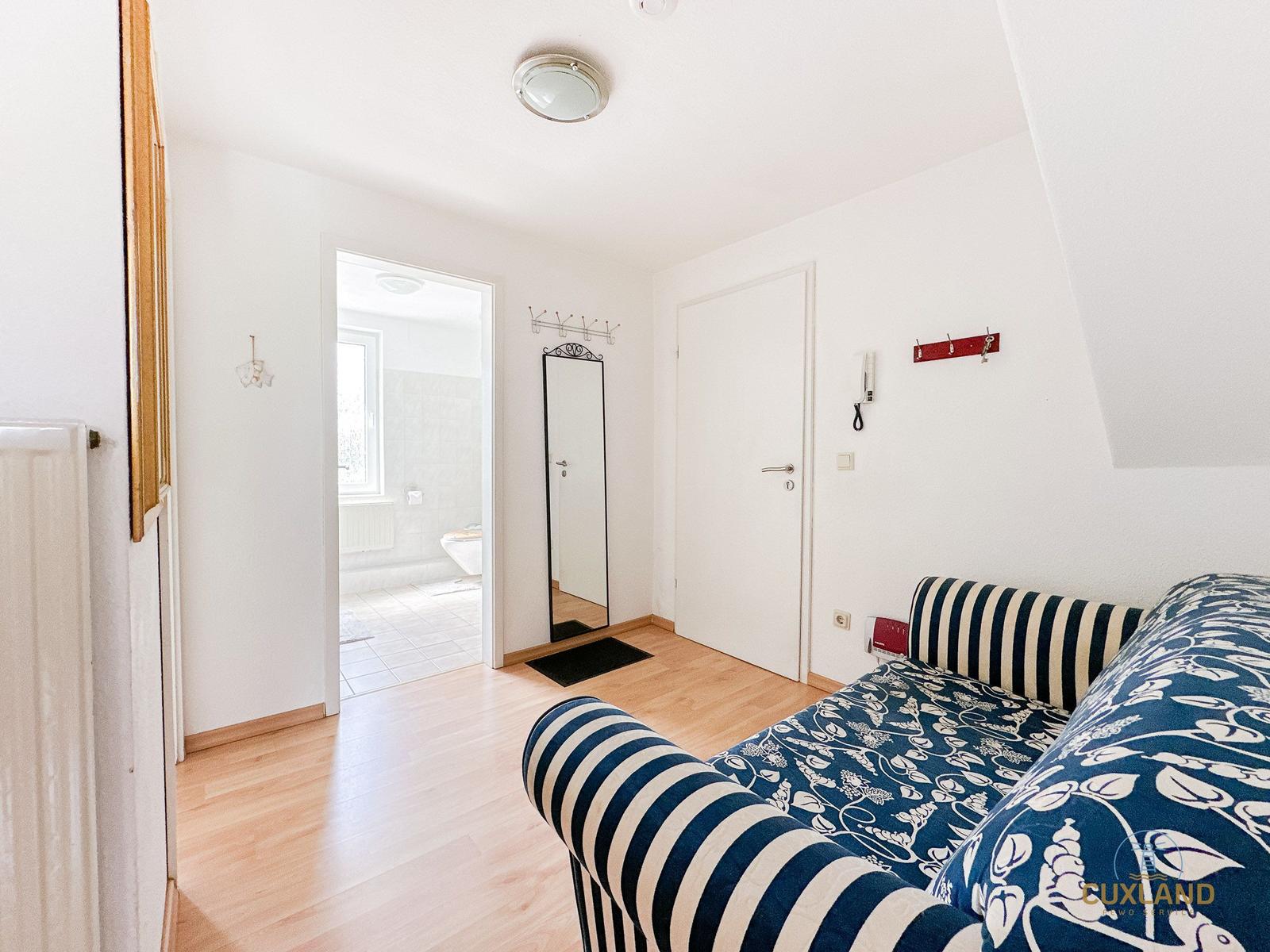 Zimmer mit Sofa, Spiegel und Tür zum Bad. Holzboden und weiße Wände.