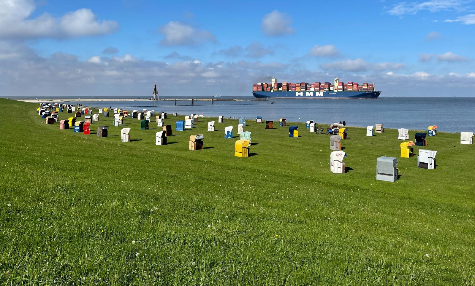 Grünes Strandufer mit bunten Strandkörben und einem Containerschiff im Hintergrund.