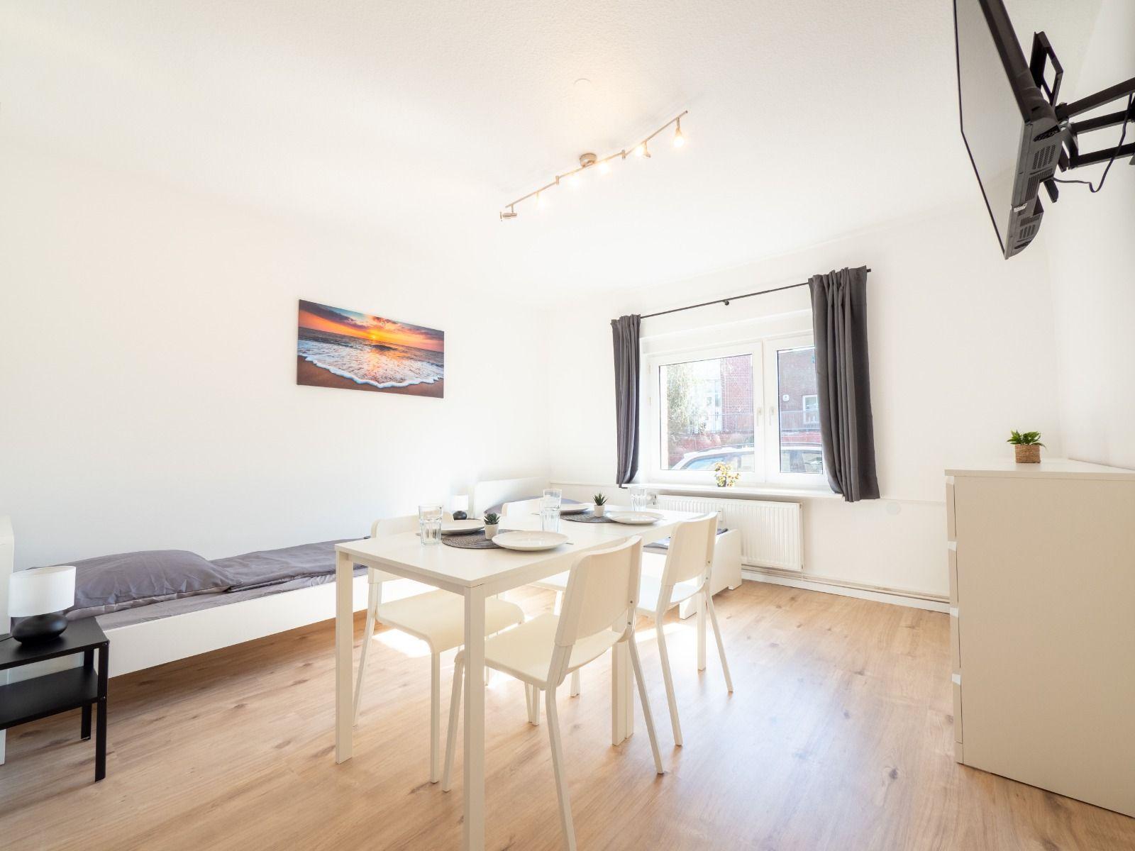 Wohnraum mit Esstisch, Bett und TV. Fenster mit Vorhängen. Wandbild und Holzboden.