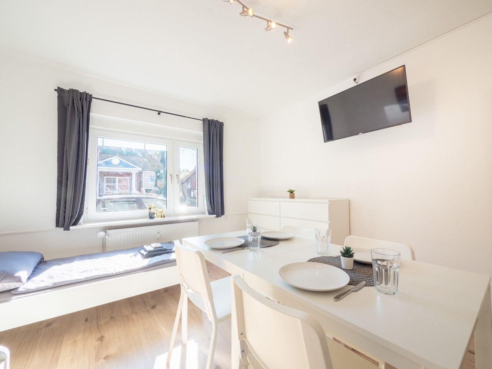 Essbereich mit Tisch, Stühlen und Fernseher. Fenster mit Vorhängen und Sitzbank.