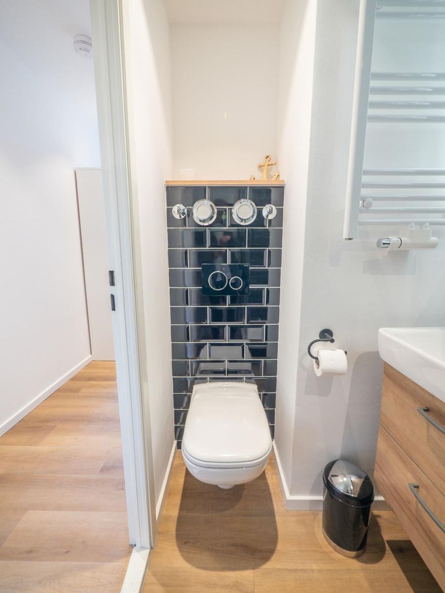 Toilette mit dunklen Fliesen und Holzboden. Toilettenpapierhalter und Abfalleimer sind vorhanden.