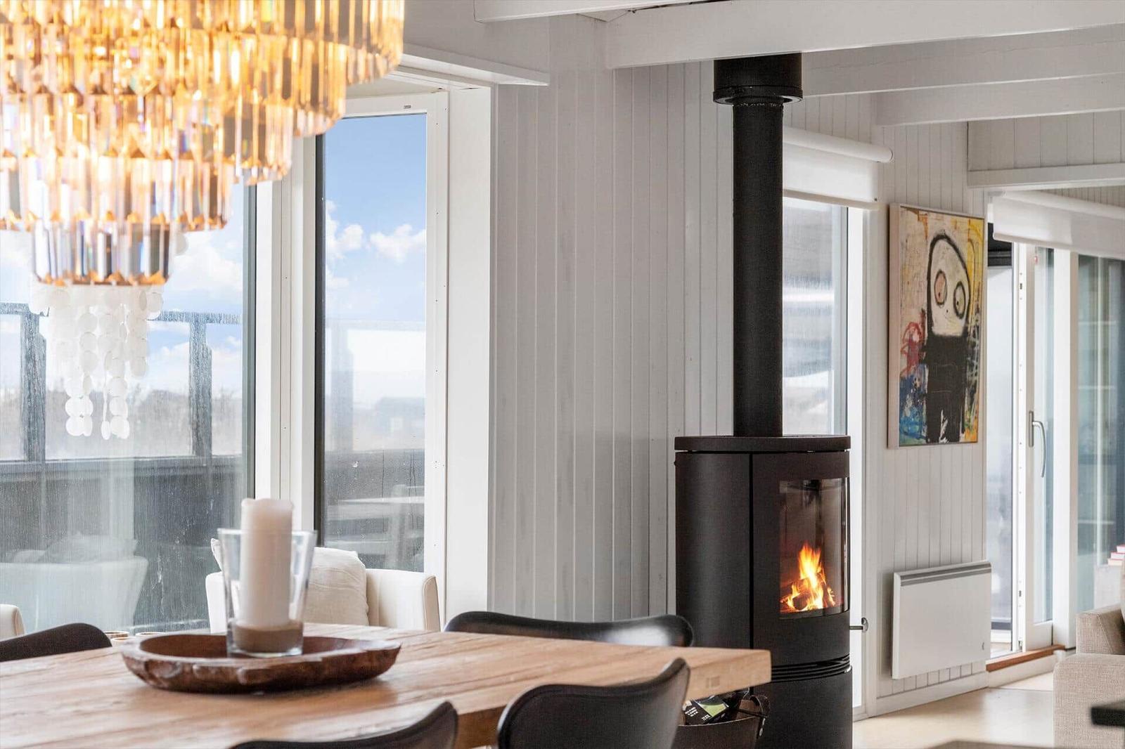 Essbereich mit Holztisch, Kaminofen und großen Fenstern. Kristallleuchte und Wandbild sichtbar.