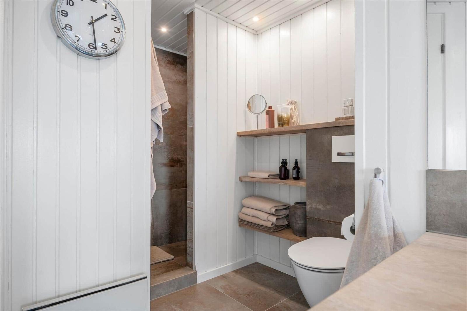 Badezimmer mit Dusche, Toilette und Holzregalen. Wanduhr und Handtücher sichtbar.