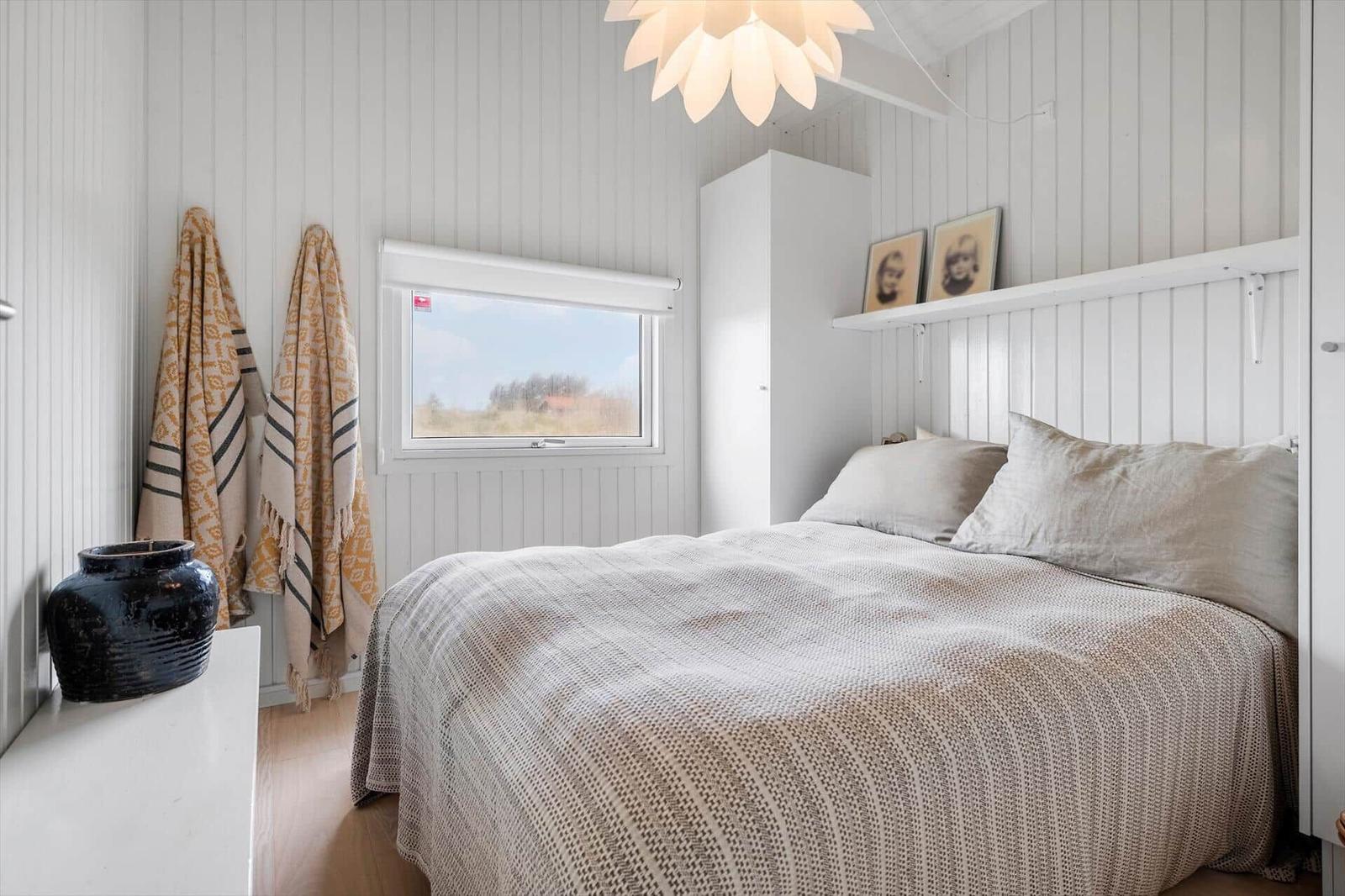 Schlafzimmer mit Bett, Fenster und Wandregal. Weiße Holzwandverkleidung und Deckenleuchte.