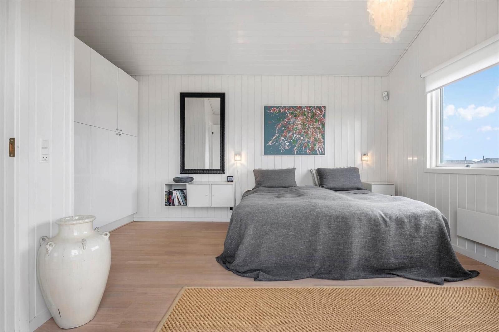 Schlafzimmer mit Bett, Wandspiegel und Fenster. Holzboden und weiße Wände.