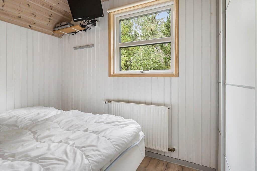 Schlafzimmer mit weißem Bett, Holzdecke und Fenster mit Blick auf Bäume.