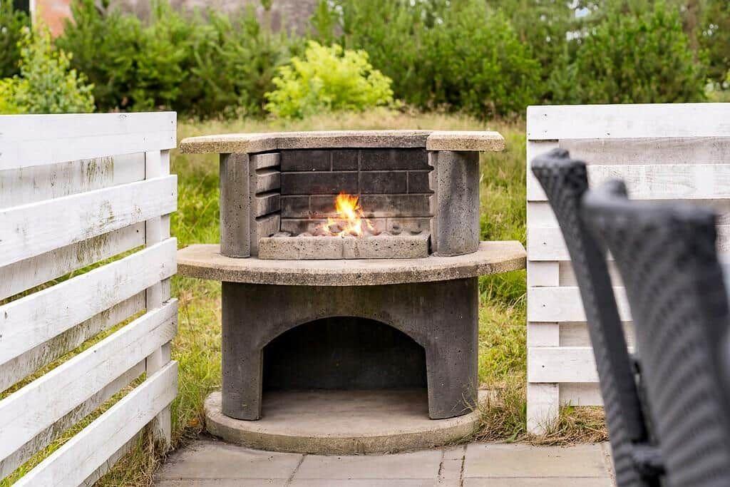 Steinofen mit brennendem Feuer im Garten, umgeben von weißen Holzpaneelen und Grünflächen.